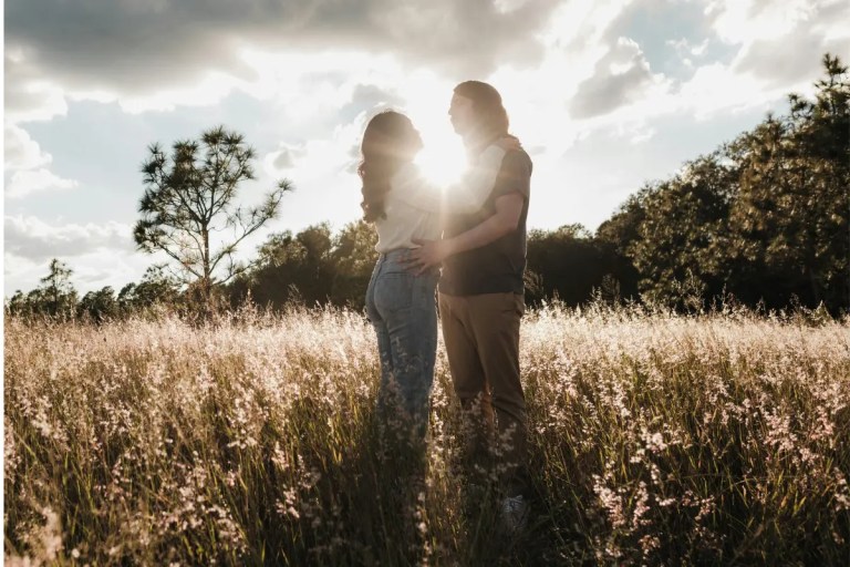 Gemini said A medium shot of a couple standing in a field of tall grass during the golden hour. The man and woman are embracing and looking at each other, backlit by the setting sun. Behind them is a treeline of pine and other trees.