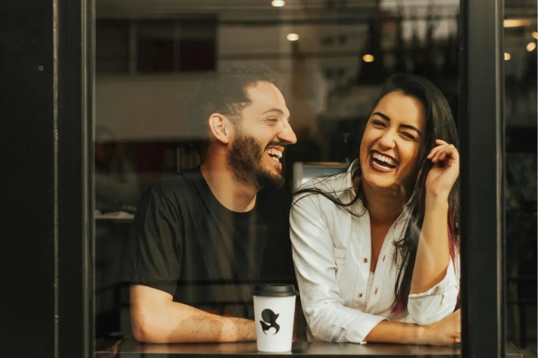 A smiling man and woman laughing together while sitting inside a cafe, viewed through a window.