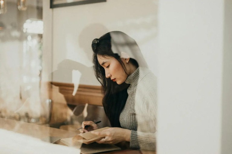 A woman in a grey turtleneck writing in a notebook inside a cafe, viewed through a glass window with a faint reflection.