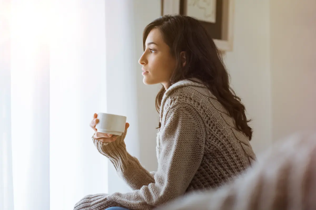 A woman in a cozy knit sweater sitting by a window, holding a white mug and looking outside