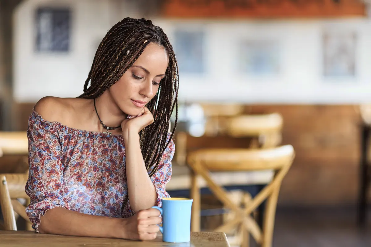 A woman with braided hair sitting at a table in a cafe, holding a blue mug and looking down pensively.