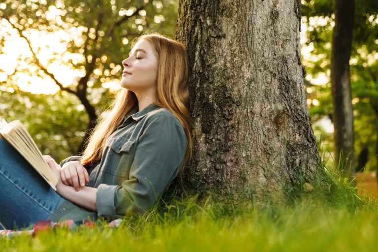 A woman rests against a tree in a sunlit park with an open book in her lap
