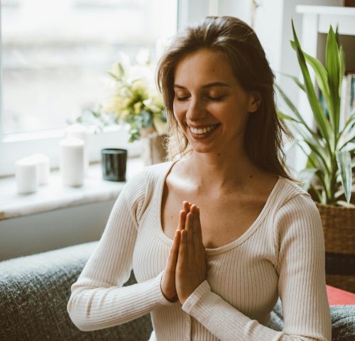 Portrait of a smiling woman meditating on a sofa with hands in prayer position.